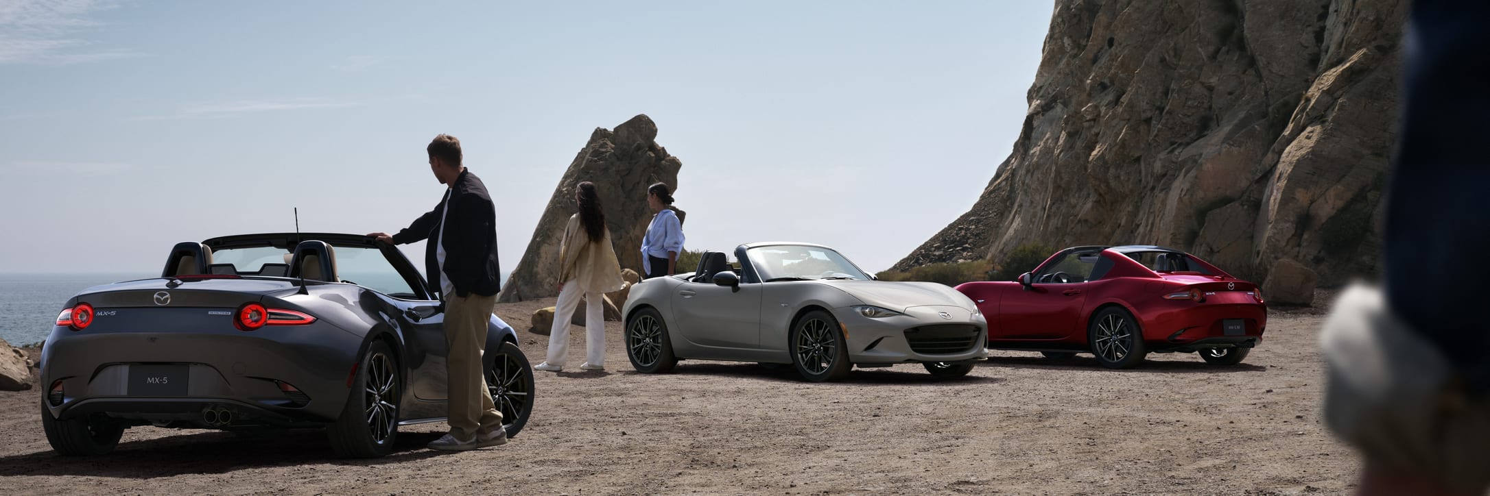 Three Mazda MX-5 roadsters on a coastal road beside rocky cliffs